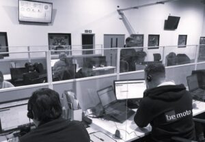 A black and white photo of a large contact centre with banks of desks. There are figures sat at the desks wearing headsets and handling customer calls. The office is decorated with pictures of motorcycles.