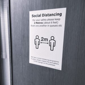 A black and white photo of a social distancing sign fixed to an office door. The sign explains the importance of keeping a suitable distance apart to limit the chance of spreading Coronavirus.