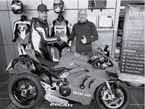 A black and white photo of a BeMoto employee and a competition winner shaking hands behind a Ducati Panigale V4S super sports bike with a "win me" sticker on the right side fairing.