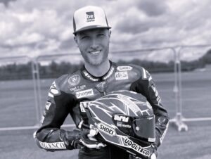 A black and white photo of Davey Todd smiling and wearing a BeMoto branded cap and motorcycle leathers, holding his race helmet.