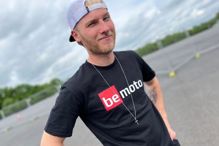 Davey Todd wearing a BeMoto branded T-shirt and cap, standing in an open area with cloudy skies and fencing in the background.