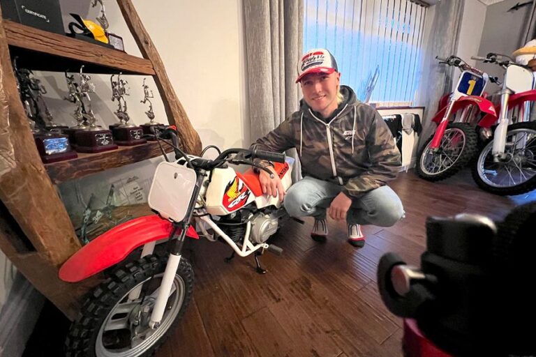 Davey Todd kneeling indoors next to a red and white Honda QR50 mini bike, with a trophy shelf and motocross bikes in the background.