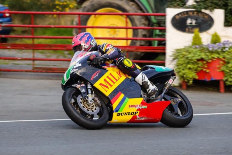 Davey Todd cornering on a Padgetts Honda RS250 race bike in full racing gear, passing a rural backdrop and red gate.
