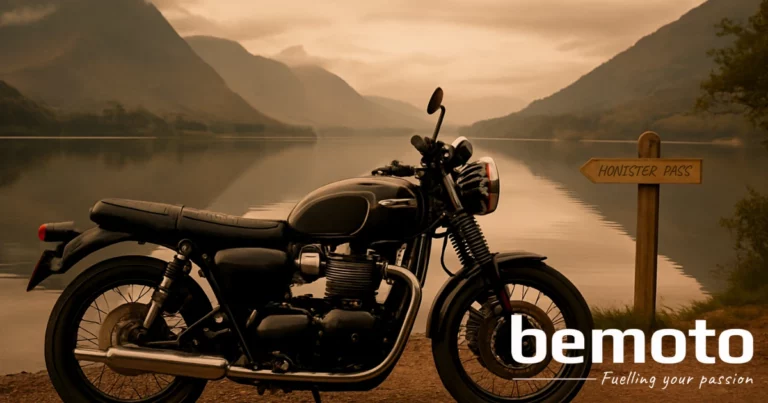 A black modern retro motorbike is parked beside a calm lake in the Lake District, with mountains and clouds reflected in the still water. A wooden signpost reads "HONISTER PASS," pointing towards the fells. The image has a warm, moody tone with an overcast sky, and the BeMoto logo with the tagline "Fuelling your passion" is placed in the bottom right corner.