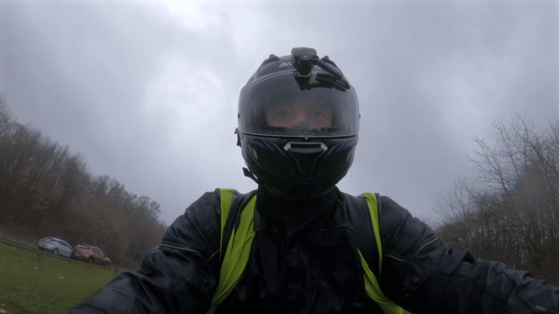 A looping animated image of a motorcyclist wearing wet weather gear and a hi vis vest, riding a motorbike in poor conditions. There is a BikerGuard visor wiper unit installed on his helmet, wiping the rain away from his visor as he rides.