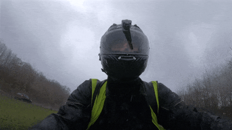 A looping animated image of a motorcyclist wearing wet weather gear and a hi vis vest, riding a motorbike in poor conditions. There is a BikerGuard visor wiper unit installed on his helmet, wiping the rain away from his visor as he rides.