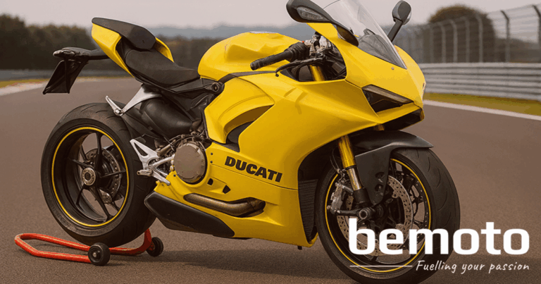 A bright yellow Ducati Panigale sports bike is parked on a racetrack paddock stand, with black and yellow wheels and visible gold front forks. The background shows track fencing and blurred trees. The BeMoto logo sits in the lower right corner.