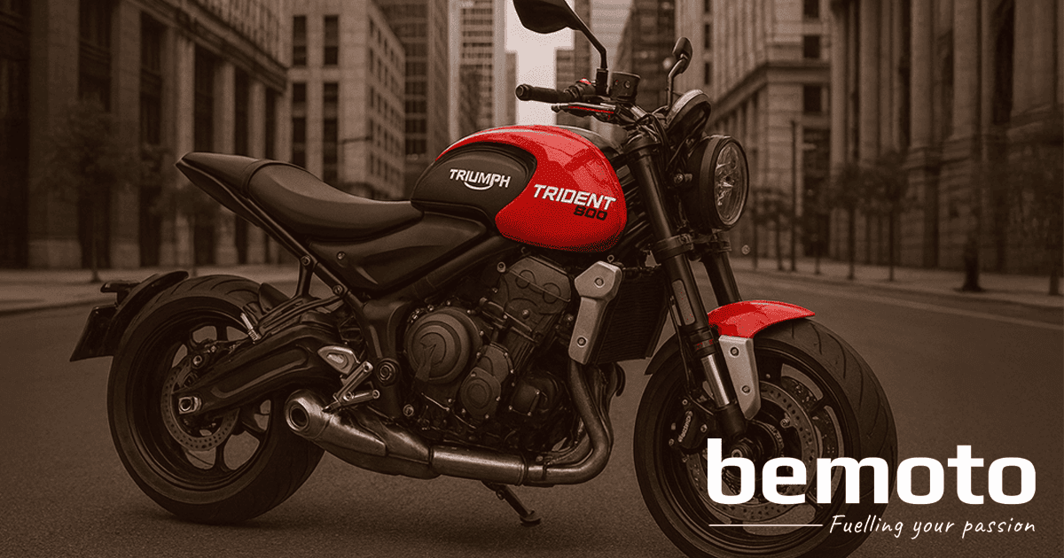 A red and black Triumph Trident street bike is parked on an empty city road, surrounded by tall buildings with a mix of modern glass and older stone architecture. The bike stands upright without a paddock stand, and the urban setting highlights its naked, sporty design.
