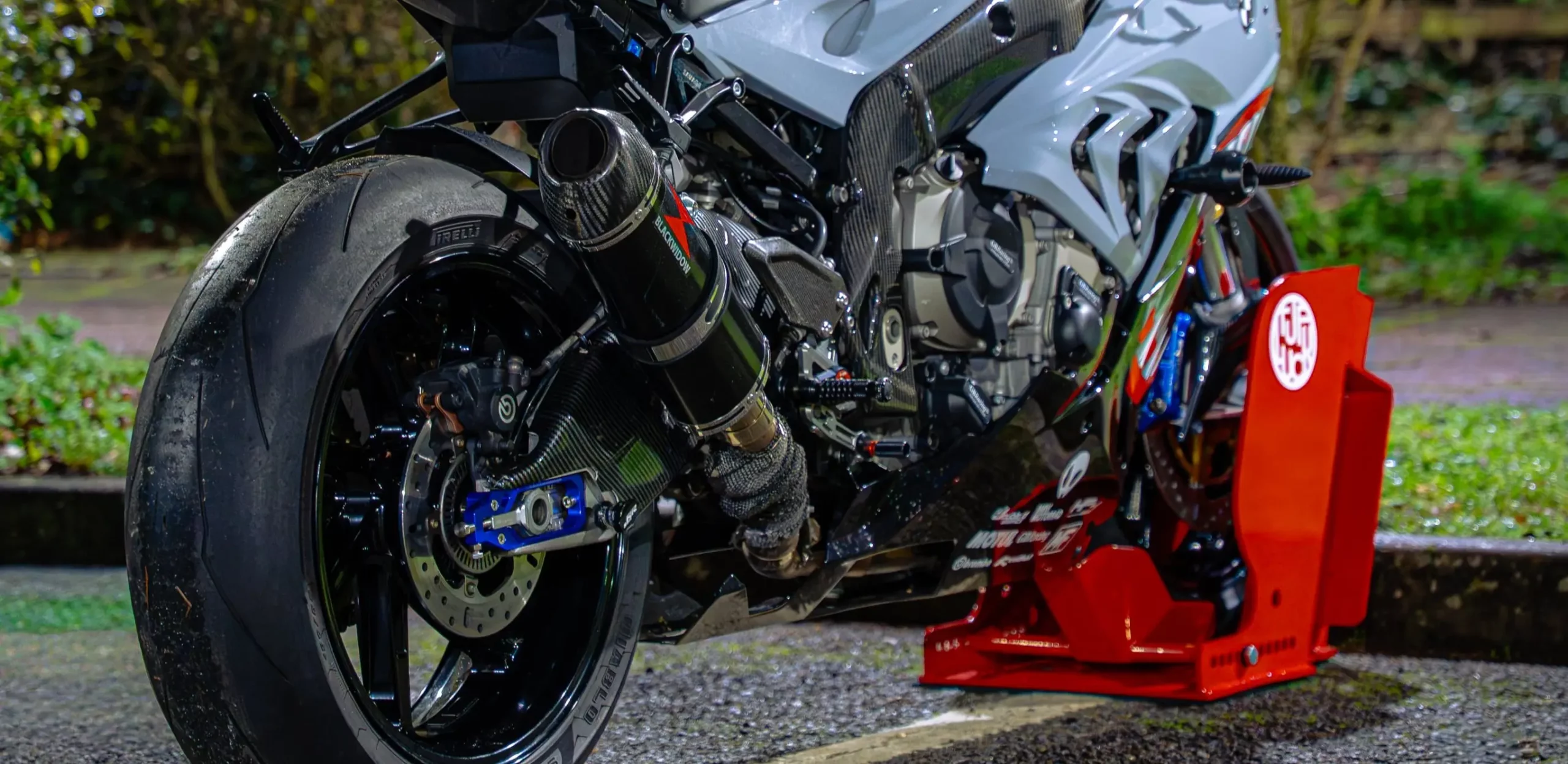 Rear three-quarter view of a high-performance BMW sports motorcycle secured with a red TufLuck Tough Motorcycle Parking Lock, showing the rear wheel, exhaust, and integrated ground anchor.