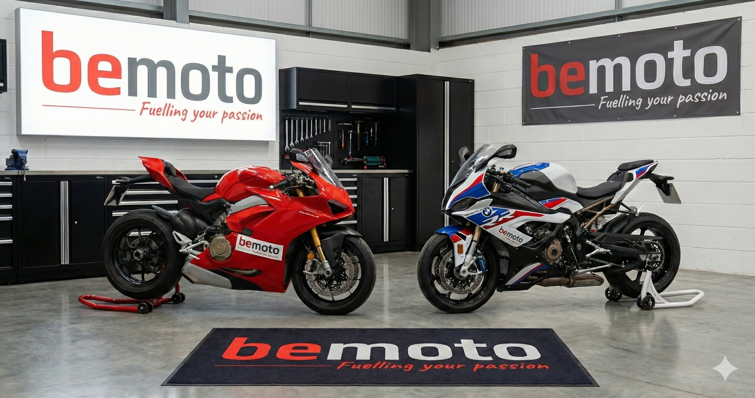 Red Ducati 1199 Panigale and BMW S1000RR motorcycles on paddock stands in a garage with prominent BeMoto Future Classics branding on a lightbox, banner, and floor mat.