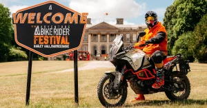 A motorcyclist in orange gear on a KTM adventure bike posing next to the official Welcome to the Adventure Bike Rider Festival sign at Ragley Hall, Warwickshire.