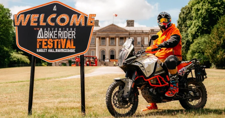 A motorcyclist in orange gear on a KTM adventure bike posing next to the official Welcome to the Adventure Bike Rider Festival sign at Ragley Hall, Warwickshire.