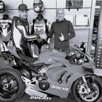 About BeMoto – Ducati Panigale V4S Giveaway A black and white photo of a BeMoto employee and a competition winner shaking hands behind a Ducati Panigale V4S super sports bike with a "win me" sticker on the right side fairing.