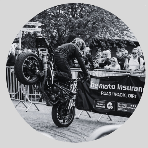 About BeMoto – MCN Festival of Motorcycling A black and white photo of a stunt rider on a motorcycle braking harshly on the front wheel, lifting the rear in the air in front of a crowd of spectators.