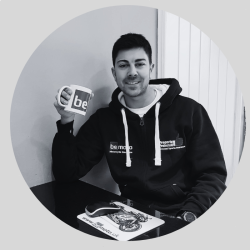 About BeMoto – Gino Rea A black and white photo of Gino Rea, smiling and seated at a desk wearing a BeMoto branded hoodie, holding a BeMoto branded mug.