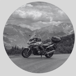 About BeMoto – Travel Insurance A black and white photo of a touring motorcycle leant over on its side stand with a beautiful mountain range in the background, surrounded by forest.